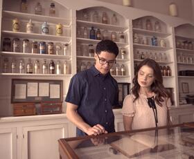 Two people examine medicinal artifacts at the New Orleans Pharmacy Museum, with a wall of bottles of various sizes and shapes behind them.