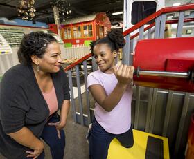 A smiling girl turns a large red crank while an adult woman watches and smiles at a colorful children's museum exhibit.