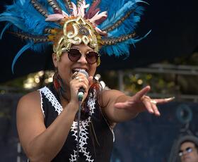 A festival singer wearing a blue feathered headdress performs on stage, singing into a microphone and reaching out toward the crowd.