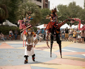 Stilt walkers and costumed performers entertain a crowd gathered in a public plaza during an outdoor festival.