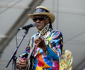 A blues guitarist in a colorful jacket sings into a microphone while playing on an outdoor festival stage.