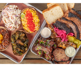 A tray overflowing with smoked meats, pickles, sliced bread and colorful sides showcases a hearty barbecue meal.
