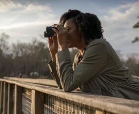 A person leans on a wooden railing while looking through binoculars over a wetland area.