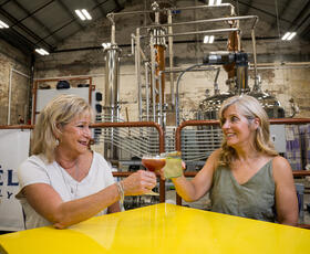 Two women raise cocktails in a cheerful toast inside the industrial-style Noël Family Distillery tasting room.