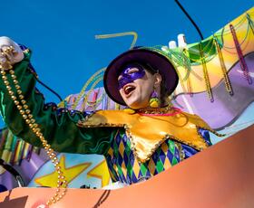 A masked parade rider tosses beads from a colorful Mardi Gras float under a clear blue sky.