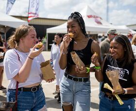 Three women laugh while eating fried festival food from paper bags at a crowded outdoor event.