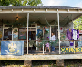 A rustic Cajun shop with porch displays of local goods welcomes a woman and child exploring its charming, weathered exterior.