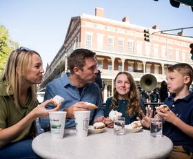 A family sits at an outdoor table at Café du Monde, eating powdered sugar–covered beignets with coffee, with French Quarter buildings and street activity behind them.