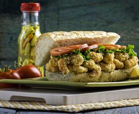 A shrimp po’boy sandwich filled with fried shrimp, lettuce, and tomato on French bread, served on a plate with pickled peppers, tomatoes, and a lemon wedge.