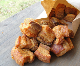 Freshly fried cracklins spilling out of a paper bag on a wooden picnic table.