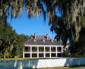 Front view of the Destrehan Plantation main house with white columns and moss-draped trees, surrounded by a white wooden fence.
