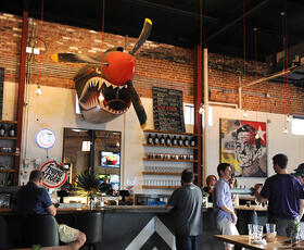 Interior of Flying Tiger Brewery in Monroe, Louisiana, featuring airplane-themed shark sculpture mounted above the bar and customers ordering drinks.