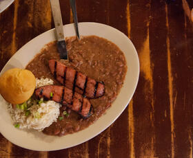 A plate of red beans, topped with sausage scored with grill marks, rice and a piece of cornbread.