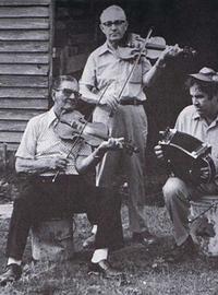 Three men play instruments — one standing with a fiddle, one sitting with a fiddle and one sitting on a white bucket with a drum