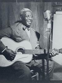 Leadbelly sits on a stool, strumming a guitar, while singing into a standing microphone