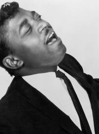 Percy, in a black suit and white shirt, eyes closed, belts out a tune with his arms in the air