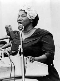 Mahalia Jackson sings from a podium on a stage. She wears a dark dress and has a fancy white hat on
