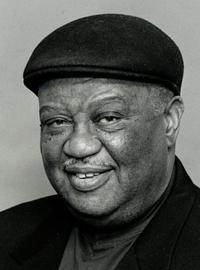 Portrait of Alvin Batiste wearing a dark hat and a jacket with a jaunty kerchief 