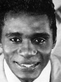 Eddie, in a sharp, light-colored suit and tie, stares into the camera. A guitar is on a backdrop behind him.