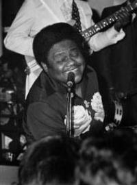 Fats sings into a microphone with a big smile on his face while playing the piano. A band plays behind him. 