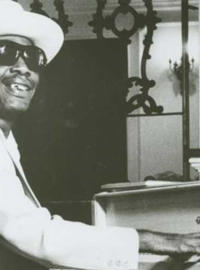 Longhair, in a light-colored suit, straw hat and sunglasses, plays a white piano