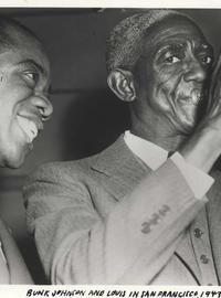 A smiling Louis Armstrong stands close to Bunk, who's playing his trumpet