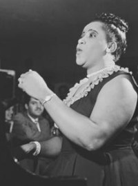 Nellie, in pearls and a black dress, hair in a bun, sings before a gathered audience