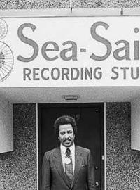 Allen, in a black suit and tie, stands in front of the entrance to Sea-Saint Recording Studio. The sign for the studio features a swirling piano keyboard and two large music notes