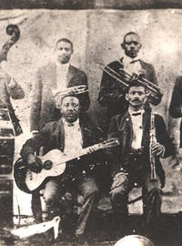 Buddy Bolder and his bandmates; four men stand with their instruments, and two sit; we see a base, guitars, trombones and clarinets