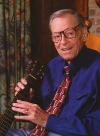 A color photo of Jimmie holding a guitar while sitting in a rocking chair. Curtains and a brick wall are behind him. He wears a navy dress shirt and patterned tie
