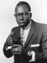 Slim points a finger at the camera in a playful way. He wears a suit with a diamond design