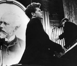A black-suited Van Cliburn sits at a piano, playing with his eyes closed. A portrait of Tchaikovsky is propped behind him