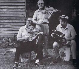 Three men play instruments — one standing with a fiddle, one sitting with a fiddle and one sitting on a white bucket with a drum