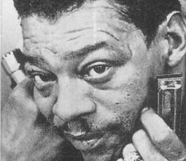Little Walter in an extreme closeup of his phase. He wears a blingy ring and holds a harmonica in each hand near his face