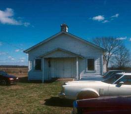 Under blue skies, a white clapboard church sits in a grassy field. 1970s cars are parked outside