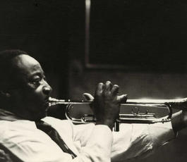 A side view of Dave Bartholomew blowing big cheeked into a trumpet in a dark room. 