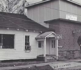 A man stands in front of two buildings, one a white clapboard house with a sign that says "Eddie's Music House," and one that's a double story, with a sign that says "Goldband Records."