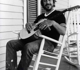 Bobby Charles sits on in a rocking chair holding a guitar on a home's front porch