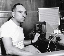 Cosimo, in a white T shirt and tweed pants, sits in front of recording equipment