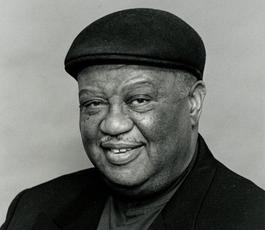 Portrait of Alvin Batiste wearing a dark hat and a jacket with a jaunty kerchief 