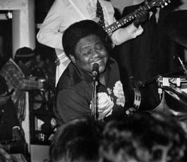Fats sings into a microphone with a big smile on his face while playing the piano. A band plays behind him. 