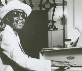 Longhair, in a light-colored suit, straw hat and sunglasses, plays a white piano