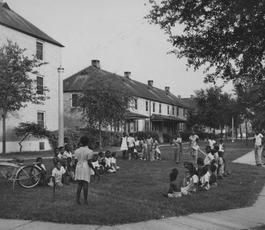 A black and white image of two housing blocks. On the lawn two dozen people lounge.