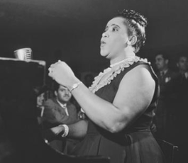Nellie, in pearls and a black dress, hair in a bun, sings before a gathered audience