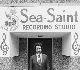 Allen, in a black suit and tie, stands in front of the entrance to Sea-Saint Recording Studio. The sign for the studio features a swirling piano keyboard and two large music notes
