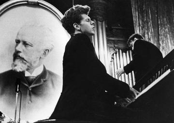 A black-suited Van Cliburn sits at a piano, playing with his eyes closed. A portrait of Tchaikovsky is propped behind him