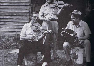 Three men play instruments — one standing with a fiddle, one sitting with a fiddle and one sitting on a white bucket with a drum