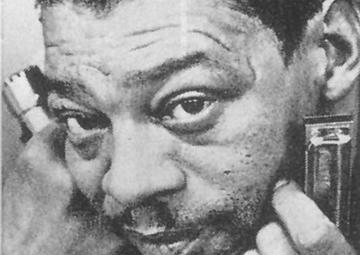 Little Walter in an extreme closeup of his phase. He wears a blingy ring and holds a harmonica in each hand near his face