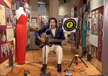 Bobby Rush is seated, playing guitar in front of a microphone in a museum. He's next to a display with a striking red ballgown and other music memorabilia
