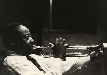 A side view of Dave Bartholomew blowing big cheeked into a trumpet in a dark room. 